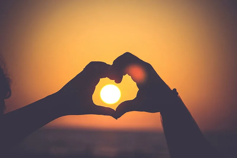 Silhuetter av to hender som former et hjerte rundt solen ved en solnedgang over havet.